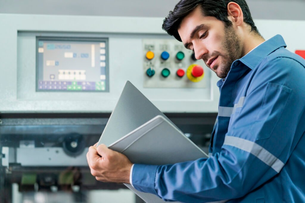Technician in industrial uniform holding technical documents in front of a machine control panel