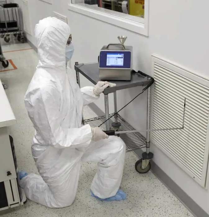 Technician in full cleanroom suit measuring air quality at a wall vent using a particle counter in a sterile lab environment.
